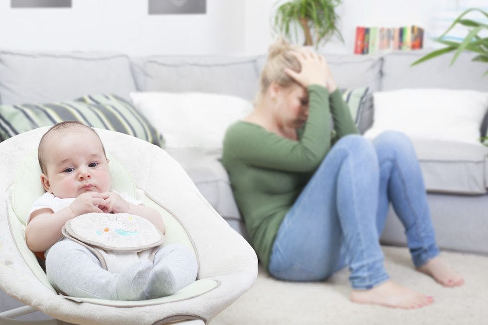 A woman sitting on the floor looking upset with her baby in a bouncer