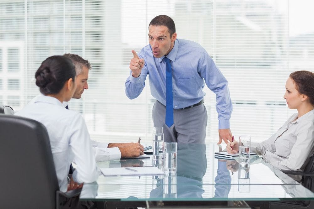 A man standing up in an office meeting pointing and yelling at coworkers