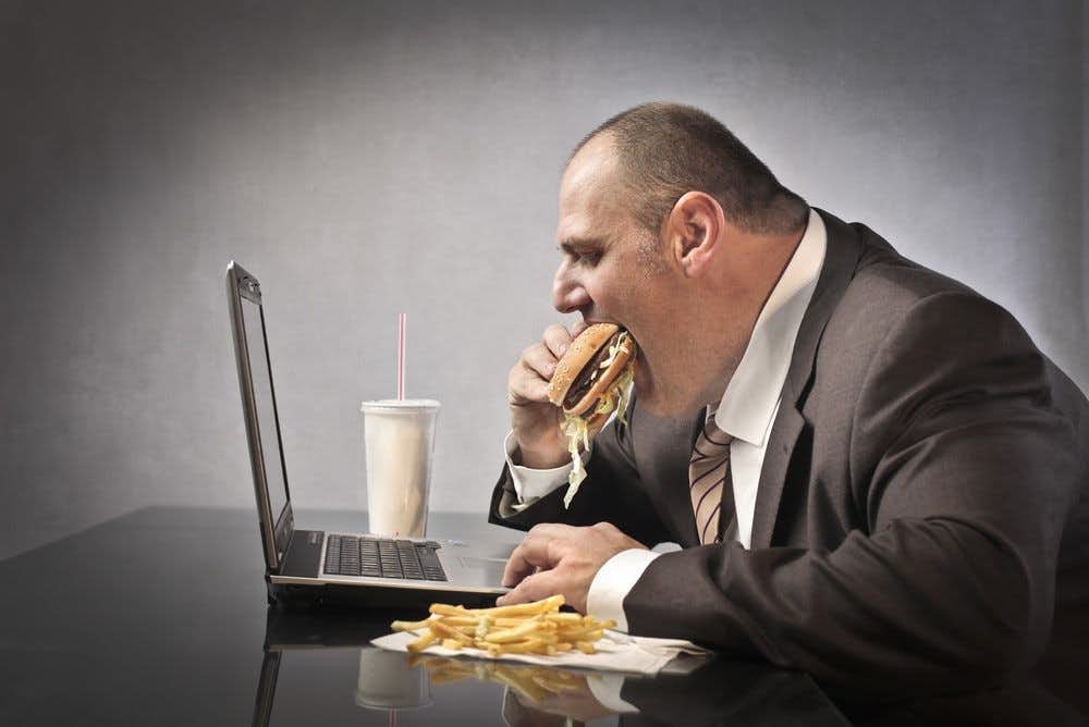 A businessman in a suit using a computer while eating a cheeseburger and fries