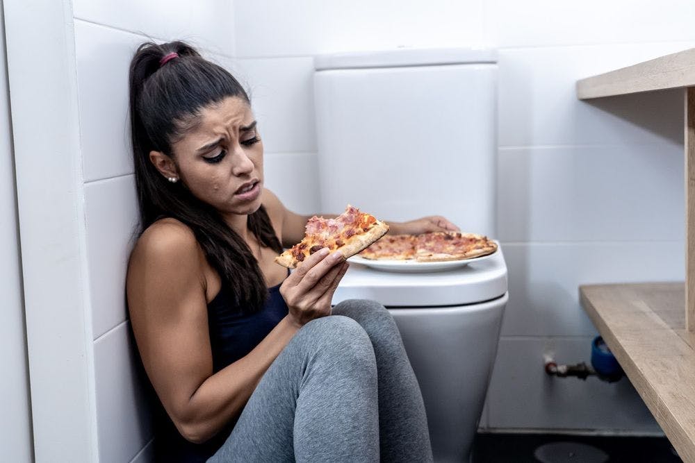 A woman eating a pizza while sitting on the bathroom floor next to the toilet
