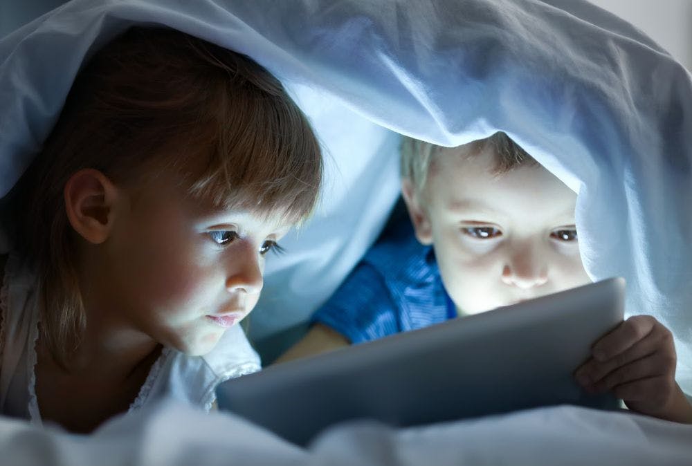 Young boy and girl under the covers looking at an ipad screen