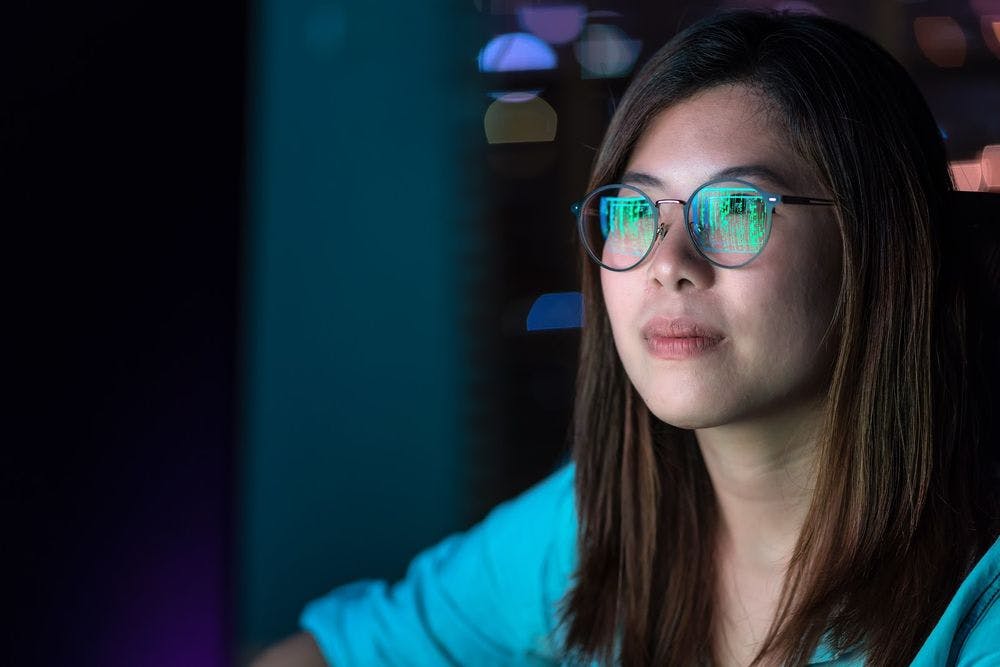 Young woman in a dark room staring at computer screen reflecting in her glasses 