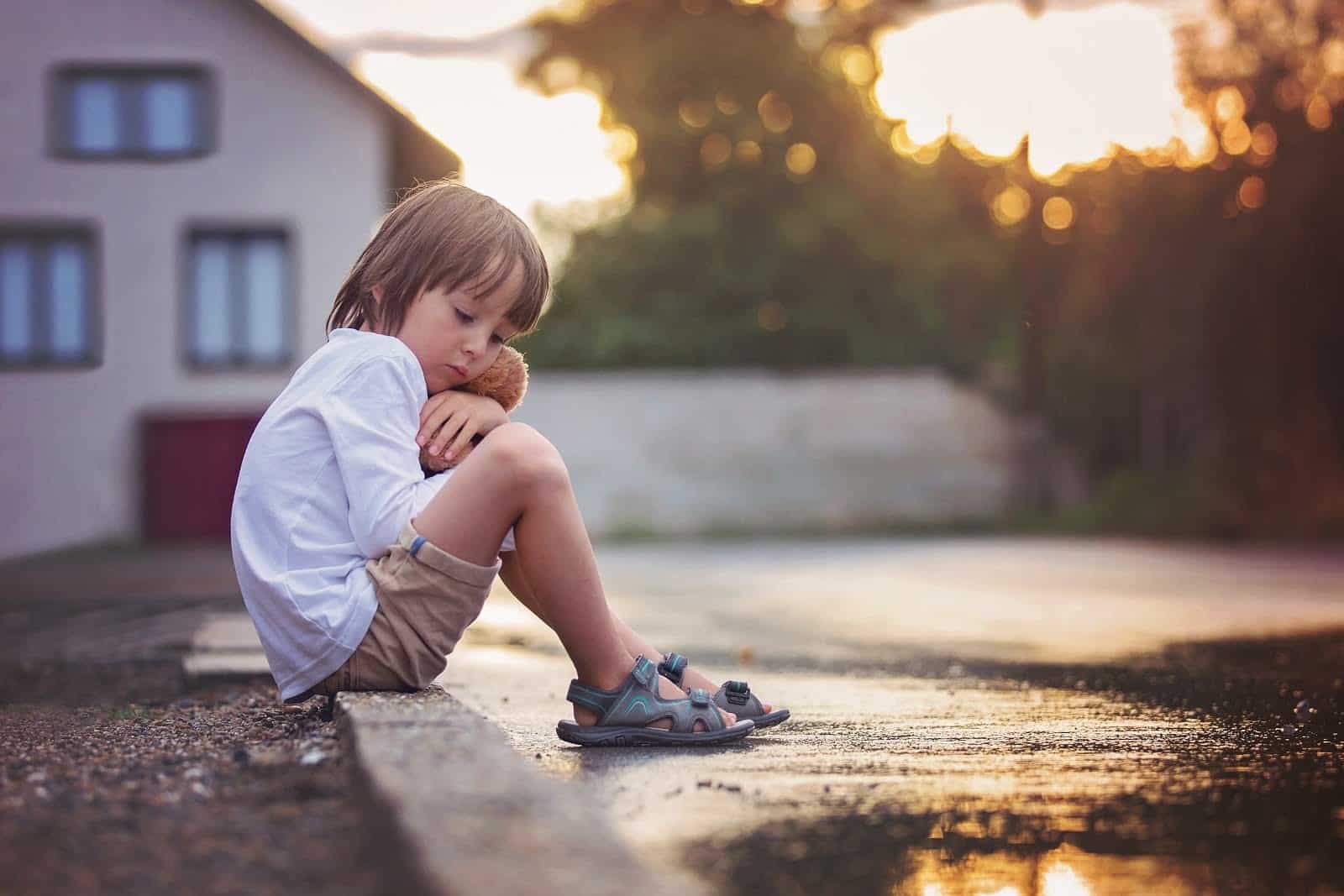 Sad little boy, sitting on the street in the rain, hugging his teddy bear, summertime on sunset
