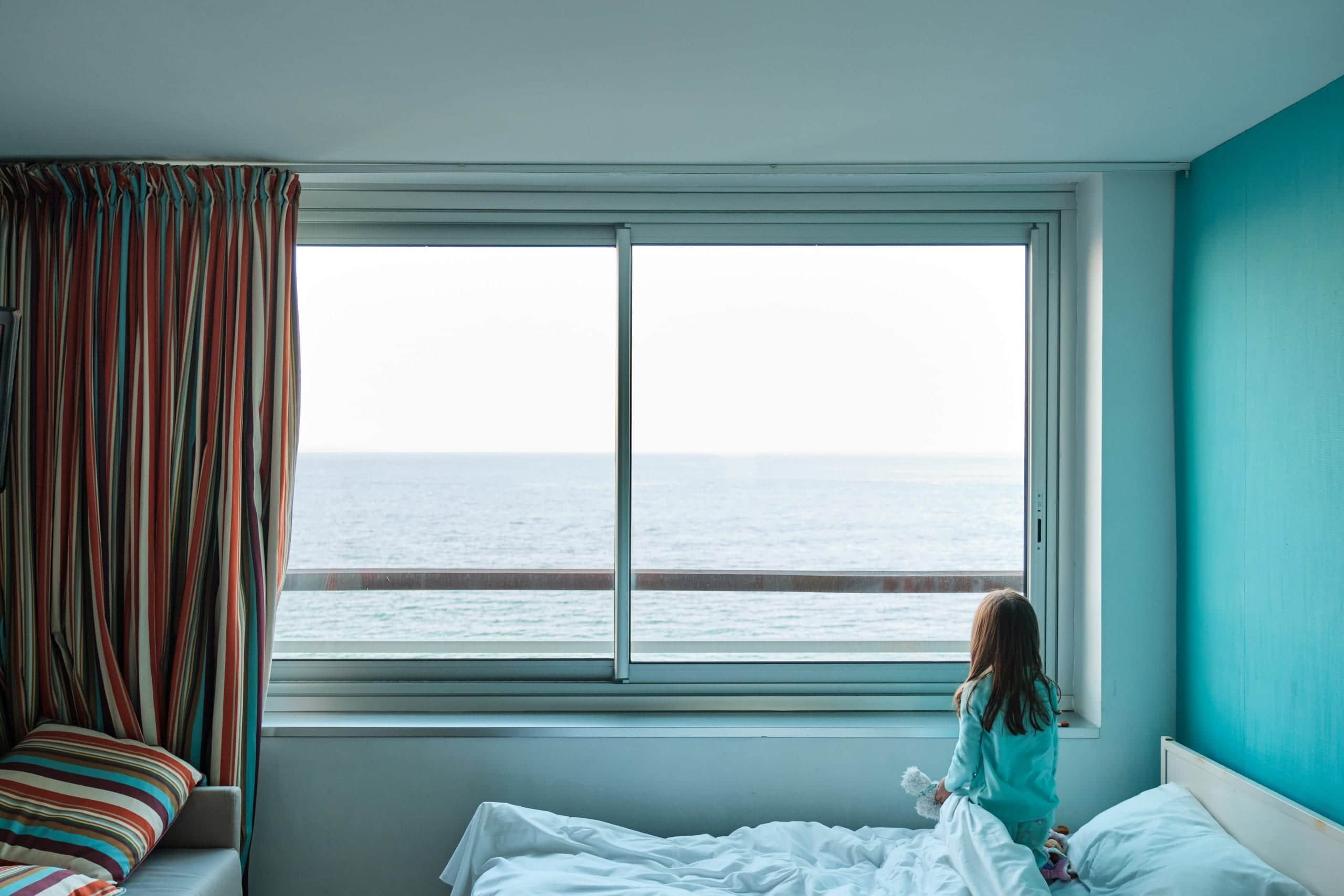 A little girl looks at the sea through the window of a hotel room